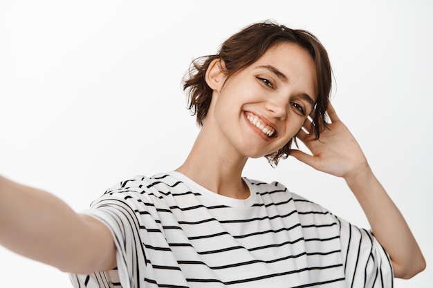 Beautiful brunette woman taking selfie, extend hand, holding camera and posing, standing in t-shirt against white background.