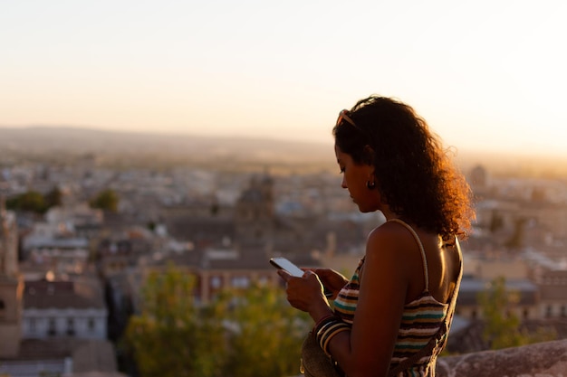 A beautiful brunette woman using her phone during sunset with the cityscape in the background The focus is on the phone screen