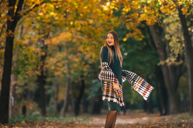 秋の自然公園で散歩を楽しんで暖かいニットのショールを身に着けている美しいブルネットの女性