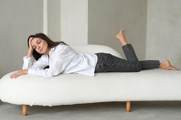 Foto bella donna castana in camicia bianca e jeans neri appoggiati su un divano bianco donna che sogna e pianifica la sua giornata