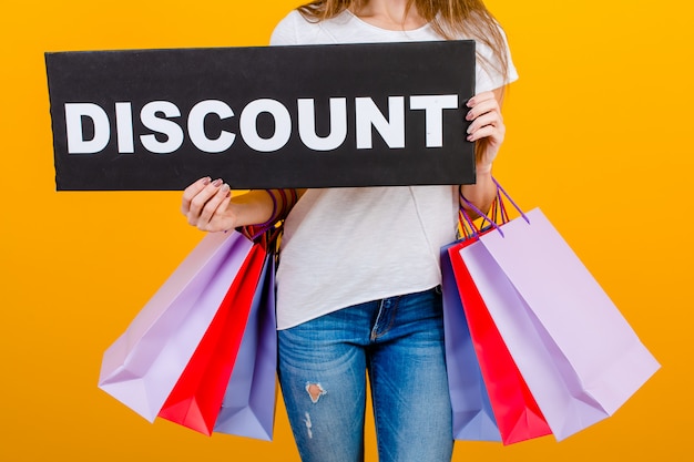 Beautiful brunette woman with colorful shopping bags and copyspace text discount sign isolated over yellow