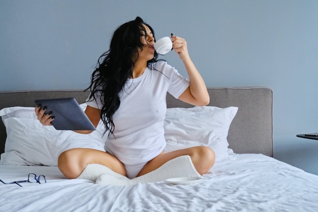 Beautiful brunette woman with cup of morning coffee sitting in bed using digital tablet, female with long wavy hair in white home clothes