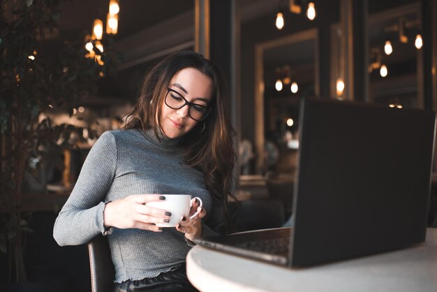 輝く光の上でラップトップに取り組んでいるカフェに座っている巻き毛の美しいブルネットの女性