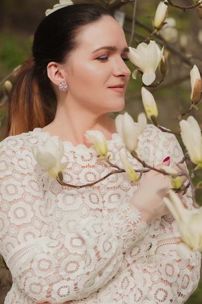 Foto bella donna bruna con trucco nudo, indossa una camicetta di pizzo, posa vicino ai fiori di magnolia in fiore