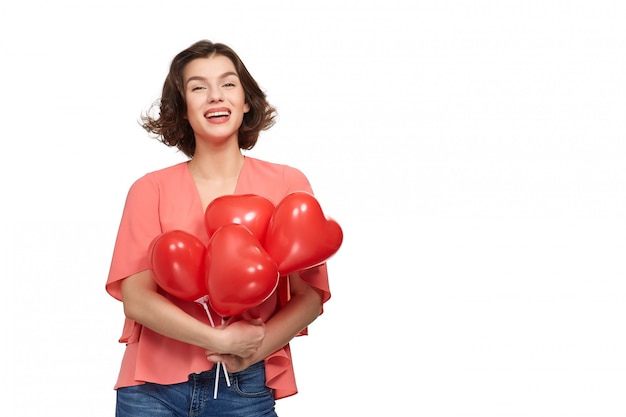 A beautiful brunette woman with a wide smile in jeans and a pink jacket  holding red balloons in the form of a heart