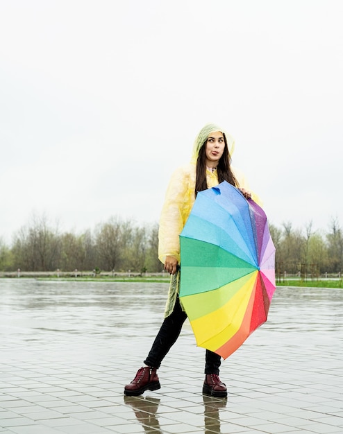 Bella donna castana in impermeabile giallo che tiene l'ombrello arcobaleno fuori sotto la pioggia, facendo una faccia buffa che mostra la lingua fuori