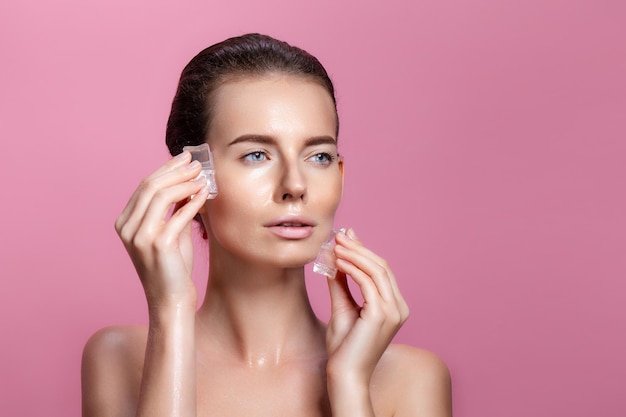 Beautiful brunette young naked female with perfect skin holds ice cubes near face on pink background