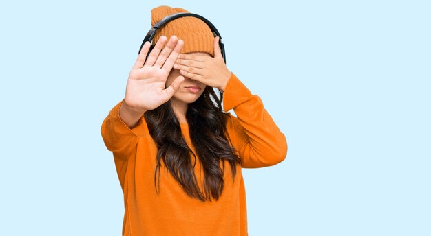 写真 美しい茶色の若い女性がヘッドフォンを使って音楽を聴いています 手で目を覆い 悲しみと恐怖の表情で ストップジェスチャーをしています 恥ずかしいし 否定的な概念です