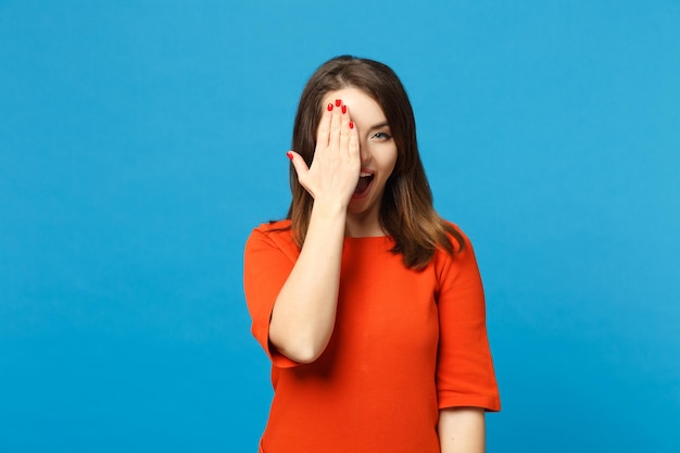 Beautiful brunette young woman wearing red orange dress hiding, covered eyes with hand palm isolated over blue wall background, studio portrait. people lifestyle fashion concept. mock up copy space