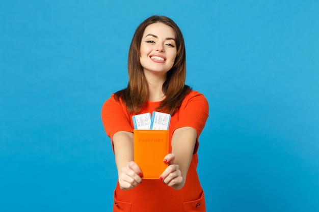 Beautiful brunette young woman wearing red orange dress hol in hand passport tickets isolated over trendy blue wall background, studio portrait. People lifestyle fashion concept. Mock up copy space.