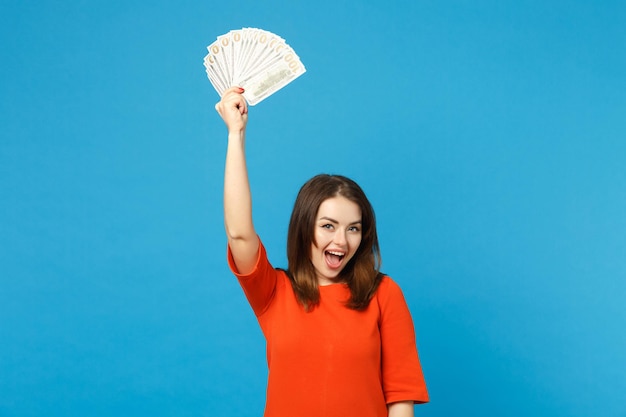 Beautiful brunette young woman wearing red orange dress holding fan of cash money in dollar banknotes isolated over blue wall background, studio portrait. People lifestyle concept. Mock up copy space.