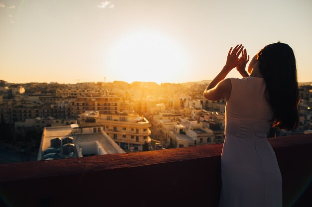 白いイブニングドレスの美しいブルネットの若い女性。街の夕日を背景に設定します。