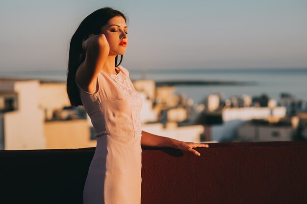 Beautiful brunette young woman in a white evening dress. setting sunset backdrop of the city. 