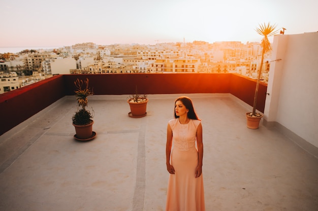 Beautiful brunette young woman in a white evening dress. setting sunset backdrop of the city. 