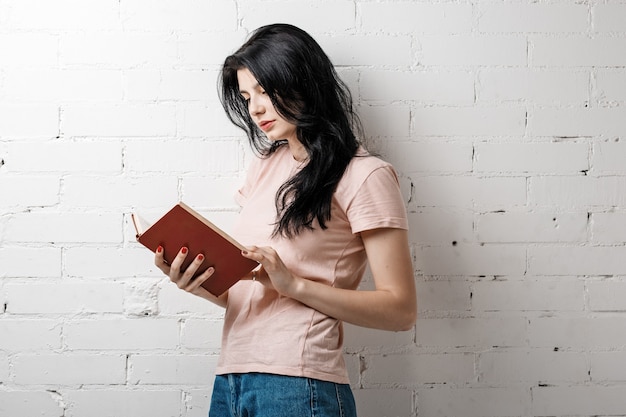 Beautiful brunette young woman with book standing near brick wall.