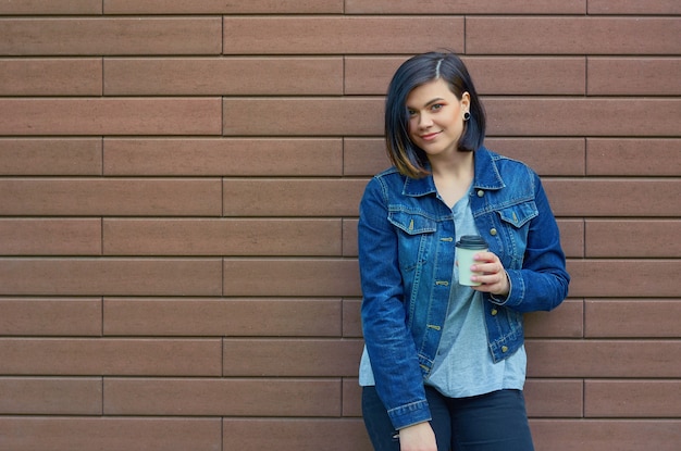 Beautiful  brunette young woman with tunnels in the ears in a blue jeans jacket  with a cup of coffee standing in front of brick wall.