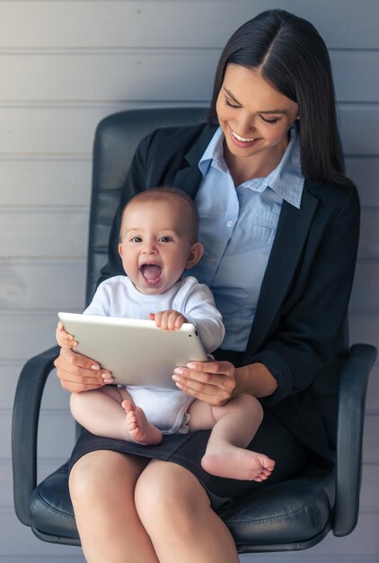 古典的なスーツを着た美しいビジネスレディと彼女のかわいい赤ちゃんは、デジタルタブレットを使用して、灰色の背景のオフィスチェアに座って一緒に時間を過ごしながら笑っています