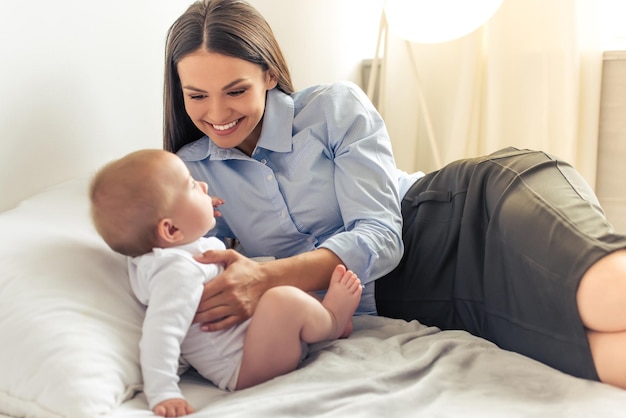 Beautiful business lady in classic suit is playing with her sweet little baby and smiling while lying on bed at home