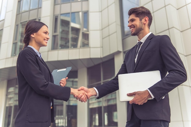 beautiful business lady and handsome young businessman in classic suits are shaking hands and smiling standing outside the office building