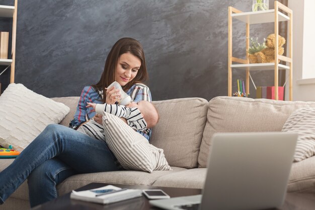 Beautiful business mom feeding child from the bottle while working on laptop in home office. Business, motherhood, multitasking and family concept.