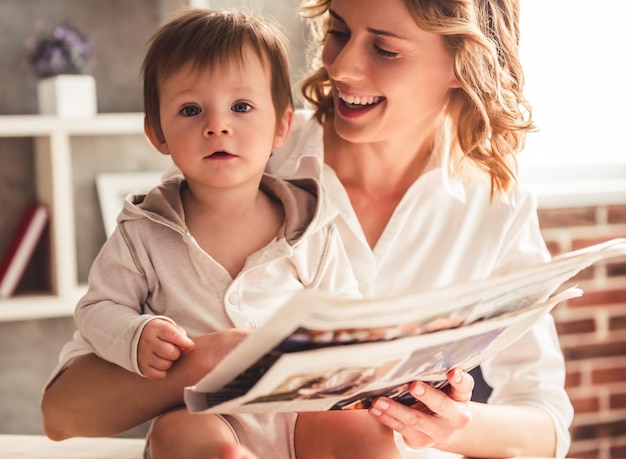 Beautiful business mom in suit is reading a newspaper with her cute baby boy and smiling at home