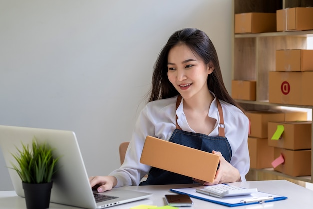 Il bellissimo imprenditore asiatico che indossa un grembiule sta preparando una cassetta dei pacchi per la consegna a un cliente utilizzando un laptop a casa.