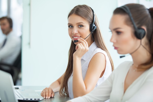 Beautiful business people in headsets are using computers and smiling while working in office. Girl is looking at camera.