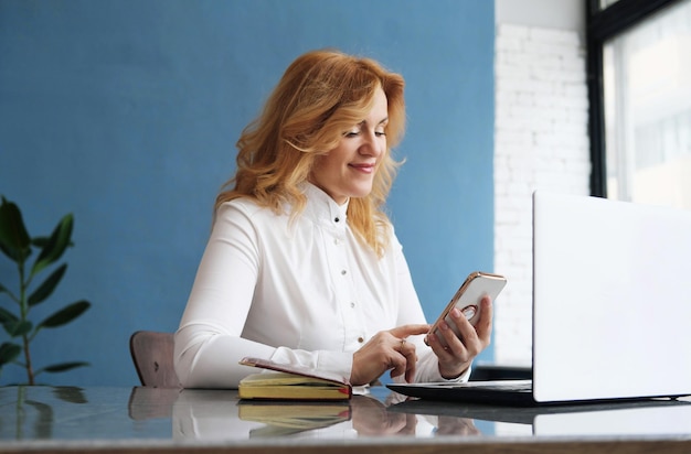 Beautiful business woman blonde in a blouse sits at a desk in\
front of an open laptop holds a mobile phone in her hands and\
answers messages