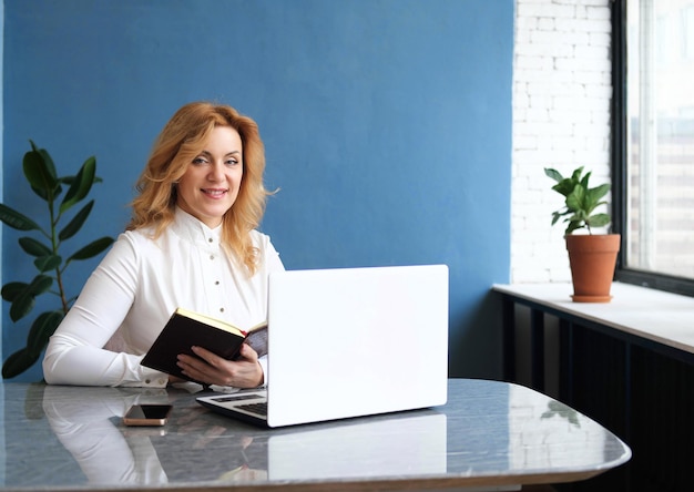 Beautiful business woman blonde in a blouse sits at a desk in\
front of an open laptop holds an open diary in her hands and smiles\
at the camera