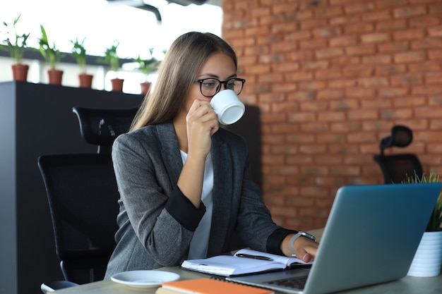 Beautiful business woman is holding coffee cup and smiling while sitting at her working place.