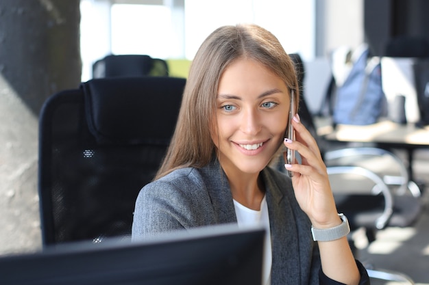 Beautiful business woman is talking on the mobile phone and smiling while sitting in modern office.