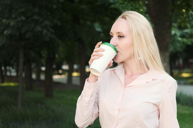 Красивая деловая женщина в рубашке, черной юбке, очках пьет кофе из одноразовой чашки, сидя на скамейке в парке, стакан для горячего напитка