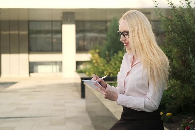 Beautiful business woman in a shirt and black skirt sits near a flower bed with a notebook and a pen in her hand Something is recording Side view