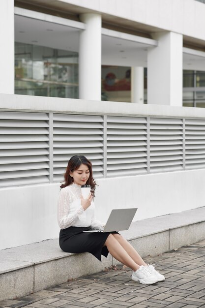 Beautiful business woman sitting on the footsteps in the financial city, using a laptop computer, thoughtful.