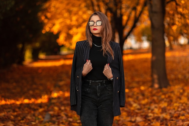 Beautiful business woman with stylish sunglasses in fashionable clothes with a blazer and a sweater walks in an autumn park with yellow fall foliage. Female elegant style and beauty