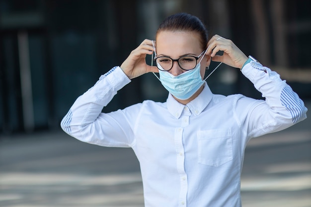 Beautiful business woman, young girl putting on medical protective mask on her face, in white shirt in glasses outdoors, healthcare at job, work, office. Coronavirus, virus, epidemic, covid-19 concept