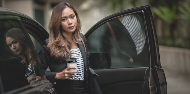 Beautiful businesswoman getting out of the modern luxury car while holding a coffee cup