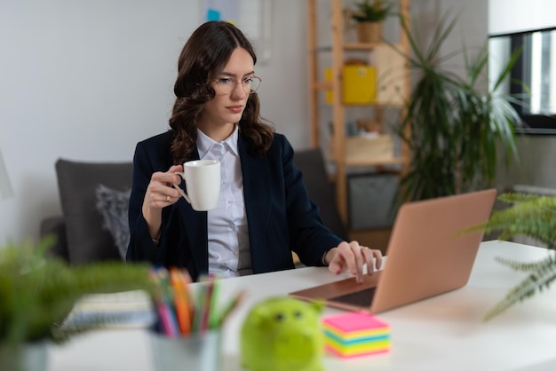 Beautiful businesswoman holding cup of coffee and looking at laptop in office