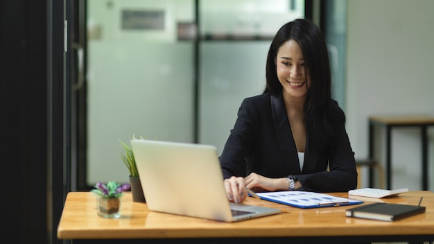 Bella donna d'affari che guarda controllando il lavoro sul computer portatile con lo spazio di lavoro dell'ufficio del viso sorridente