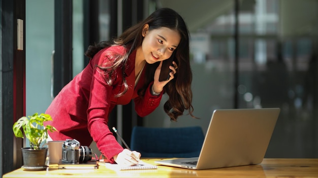 Beautiful businesswoman taking notes while having business talk on the phone with business client. Startup business concept.