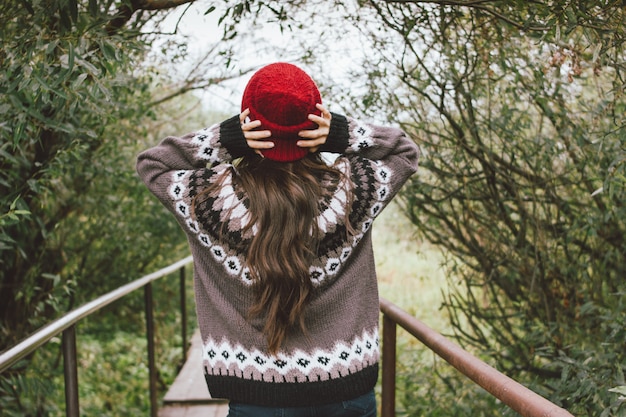 Ragazza asiatica dei bei capelli lunghi spensierati nel cappello rosso e maglione nordico tricottato da dietro nel parco naturale di autunno, stile di vita di avventura di viaggio