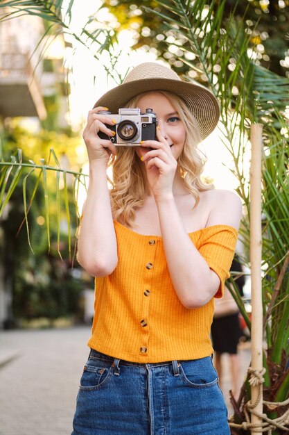 Beautiful casual blond girl in hat dreamily taking photo on retro camera on city street