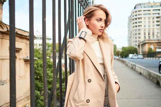 Beautiful casual blond girl in stylish trench coat thoughtfully looking aside on city bridge