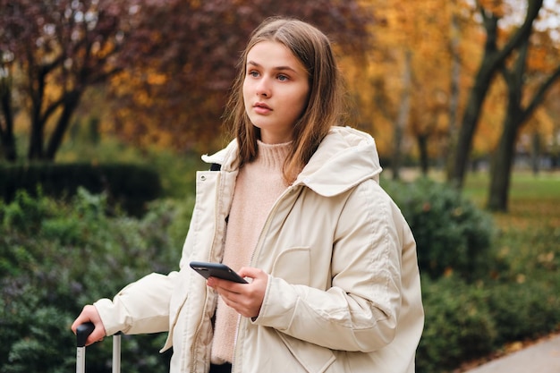 Beautiful casual girl in down jacket with suitcase and cellphone intently looking away outdoor