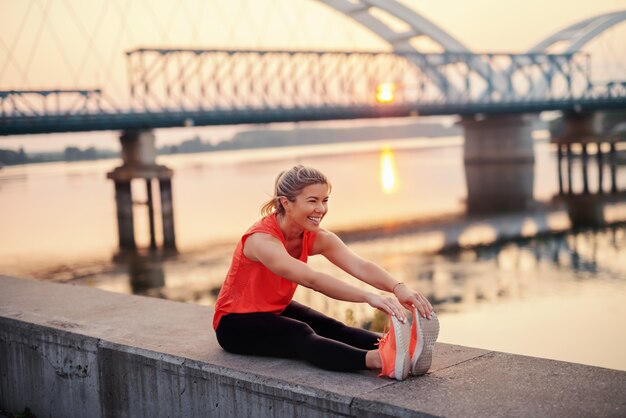 Beautiful Caucasian blonde woman in sportswear stretching while sitting on wall on quay.