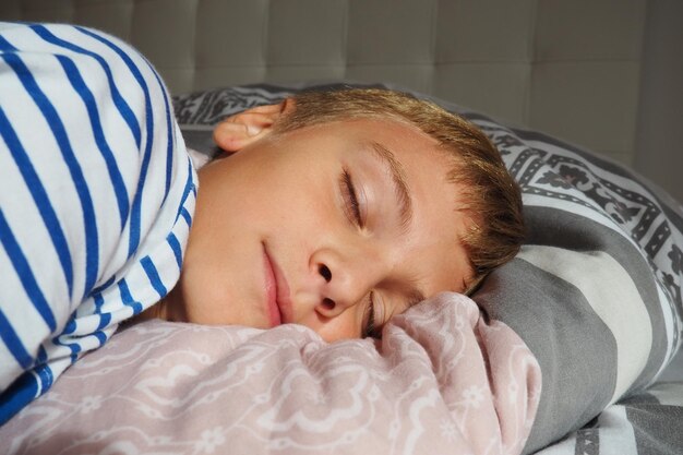 Красивый кавказский мальчик лет со светлыми волосами, одетый в полосатую пижаму, спит на кровати с