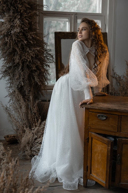 Beautiful caucasian bride girl with curly red hair stands leaning against the table in a wedding dress among dry flowers