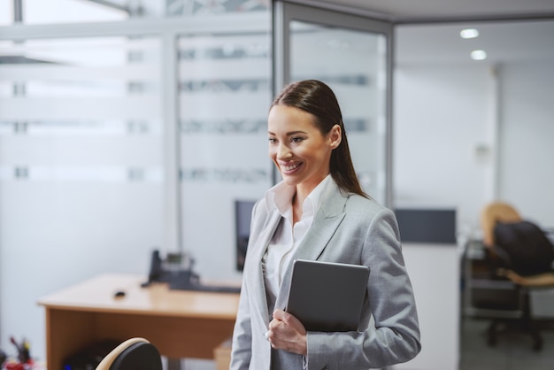 Beautiful Caucasian brunette in formal wear standing in office with tablet in hands. Great things never came from comfort zone.