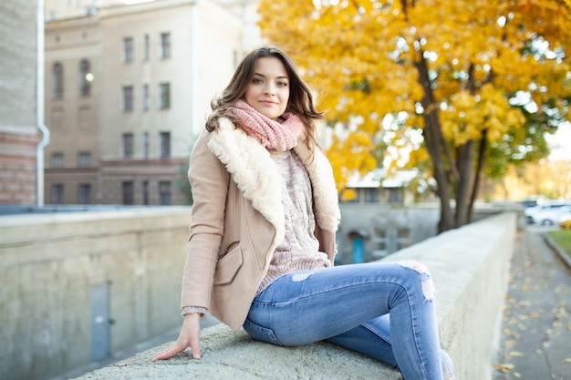 Beautiful caucasian brunette girl sitting warm autumn day with background of trees with yellow foliage and a city