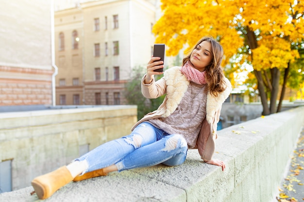 Beautiful caucasian brunette girl sitting warm autumn day with background of trees with yellow foliage and a city